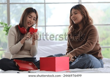 Two young Asian roommates sit on the bed and open a birthday present. They are very happy. The young friend received a gift of red headphones to listen to music in the apartment bedroom.