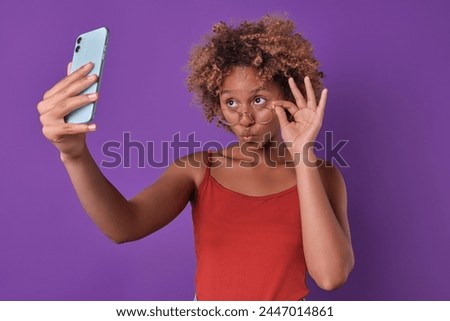 Young beautiful flirty African American woman takes selfie on phone and takes glasses off eyes posing for good photo for social networks or mobile calling applications stands in purple studio.