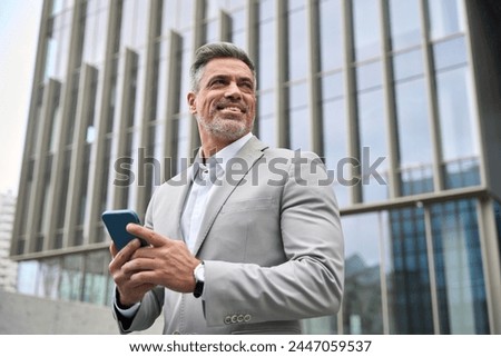 Happy business man investor of middle age in suit holding smartphone, mature professional businessman leader using mobile phone looking away standing in busy office city street with cellphone in hand.