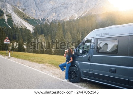 Attractive middle-aged woman leans against motorhome, sipping coffee with Dolomites peaks behind, after navigating narrow, winding roads. A quest for adventure. Royalty-Free Stock Photo #2447062345