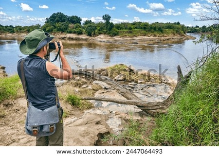 A male photographer in a green hat takes a photo the savannah on the background of river in masai mara, Kenya. The concept of an African walking safari in Africa.