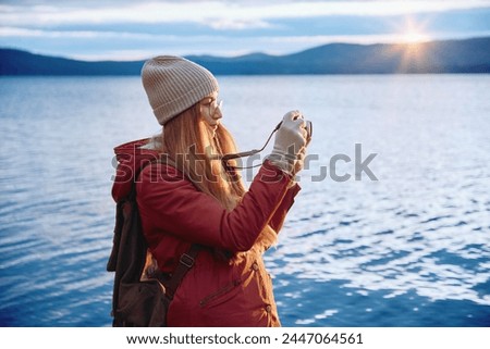 Portrait of a smiling hipster woman in glasses holding a photo camera, taking picture of a scenic lake view from cliffs. Solo traveler, explorer, freelance photographer concept