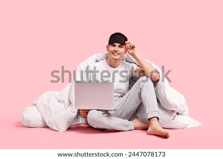Happy man in pyjama with sleep mask, blanket and laptop on pink background