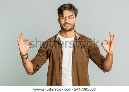 Keep calm down, relax, rest. Concentrated happy Indian man meditating breathes deeply with mudra yoga gesture, eyes closed, peaceful mind, taking a break. Arabian Hindu guy on gray studio background