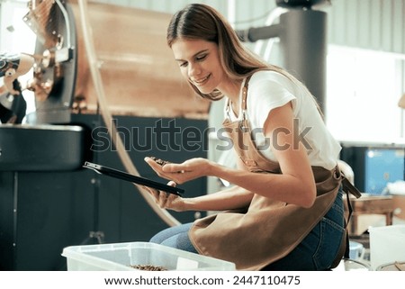 White Woman craftsperson observing and checking quality of Freshly roasted coffee beans being removed from the roaster into the plastic keeper.. Royalty-Free Stock Photo #2447110475