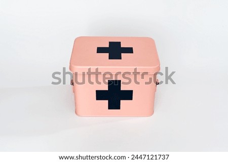 The image showcases a pink first aid box with black crosses on the top and front. It is positioned against a plain white background, emphasizing its compact and portable design. 