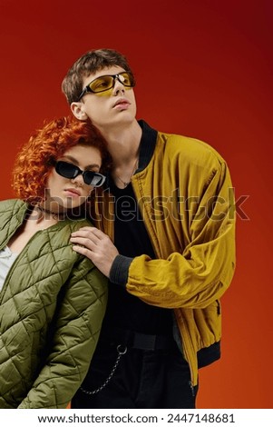 caring debonair couple in trendy sunglasses in stylish attires posing together on red backdrop Royalty-Free Stock Photo #2447148681