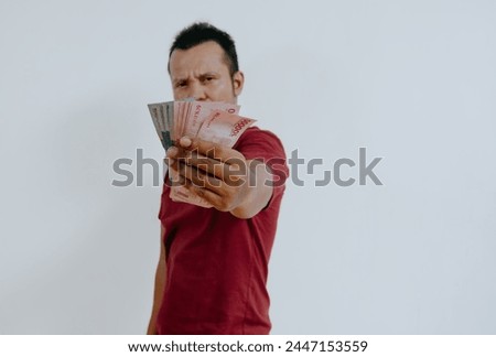 Portrait of an Indonesian Asian man, wearing a red T-shirt, showing off the money he's holding, isolated on white background.