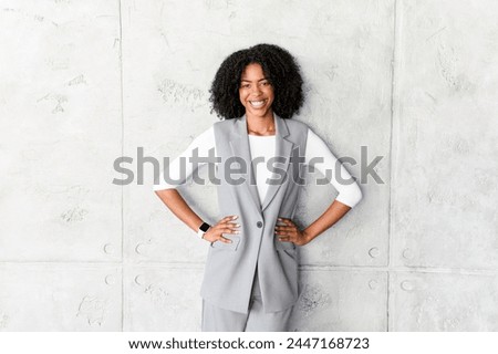 A poised African-American businesswoman stands with hands on her hips, wearing a stylish grey sleeveless vest and trousers, radiating confidence and professionalism against a textured grey background. Royalty-Free Stock Photo #2447168723