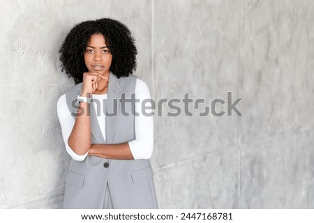 An African-American businesswoman poses thoughtfully, her chin resting on her hand, against a textured gray backdrop, symbolizing contemplation and professionalism in a corporate setting.