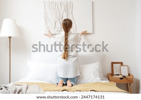 Young woman hanging 3D painting on light wall in bedroom, back view