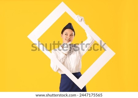 Thoughtful young Asian stewardess with frame on yellow background