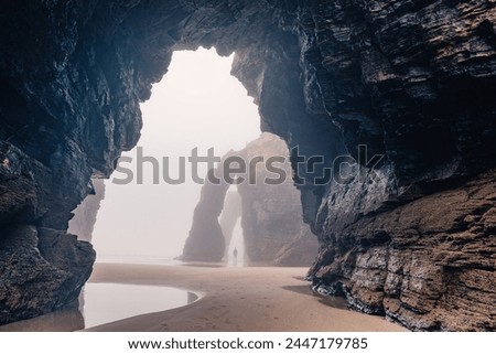 Man standing under natural arch on Cathedrals beach in Galicia, Spainn. Tourist silhouette in foggy landscape with Playa de Las Catedrales Catedrais beach in Ribadeo, Lugo on Cantabrian coast Royalty-Free Stock Photo #2447179785