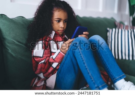 Contemplative African American girl, aged 6-8, sitting cross-legged on a green sofa, deeply engaged with a blue smartphone. She’s wearing a cozy red and white checkered shirt, blue jeans Royalty-Free Stock Photo #2447181751