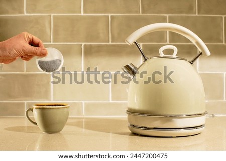 A cup of tea image. A teabag being placed into a cup. A pale green themed picture with green tiles, cup and kettle. A hand is seen holding the teabag. Hot beverage image. 