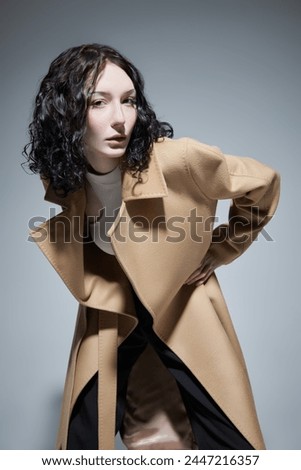 Fashion portrait of a young woman with dark wavy hair and pale skin posing in a light brown coat. Grey studio background. Makeup and cosmetics.