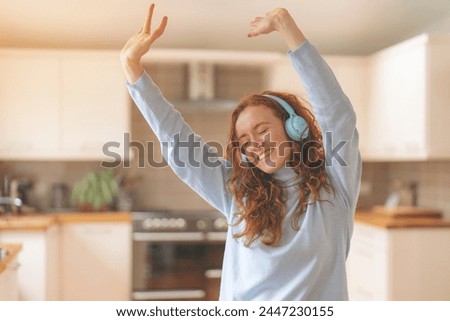 Happy young redhead woman in blue shirt using a mobile phone, listening to favorite songs, audiobooks, podcasts and dancing  on headphones while sitting in kitchen at home