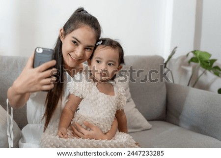 A woman and a little girl are sitting on a couch, the woman taking a picture of the girl with her cell phone. Scene is warm and loving, as the woman is capturing a special moment with her daughter