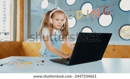 Little young cute girl using laptop to working or playing games. Student working on computer while looking and smiling at camera. Caucasian child attend online class by using headphone. Erudition.