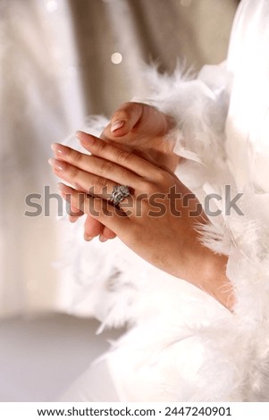 Close-up shot of a bride in a white feather dress looking at a very expensive and beautiful diamond stone ring on her hand against a dark blurred background.