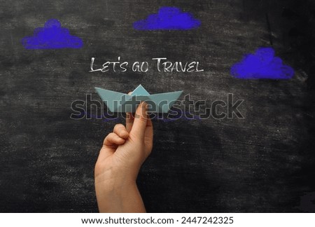 A hand holding a paper boat with the words Let's go travel written on a chalkboard