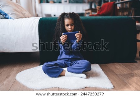 Focused African American girl, 6-8 years old, sitting cross-legged on a fluffy white rug, deeply engaged with a smartphone. She's dressed in a comfortable blue outfit, showing a blend of technology Royalty-Free Stock Photo #2447250279