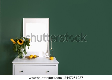 White commode in classic style with picture, vase with flowers, candles and fruits on top against green wall in cozy bedroom at home. Stylish decor elements in room. Copy space.