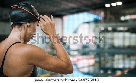 Fit female swimmer with a black swimming cap, standing looking at the pool and focusing on the coming race, putting on swimming goggles. Royalty-Free Stock Photo #2447254181