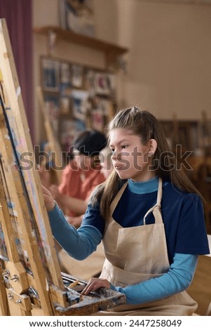 Vertical portrait of teen girl with disability painting in art class sitting by easel