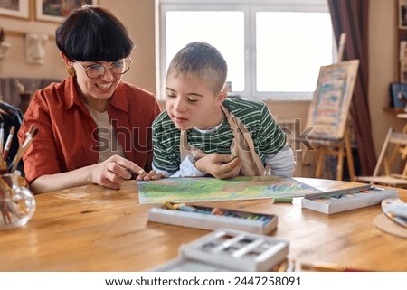 Portrait of curious young boy with Down syndrome enjoying art class in studio with woman assisting