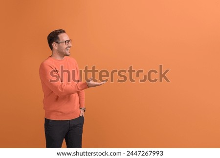 Young businessman laughing and promoting new product on empty palm over isolated orange background