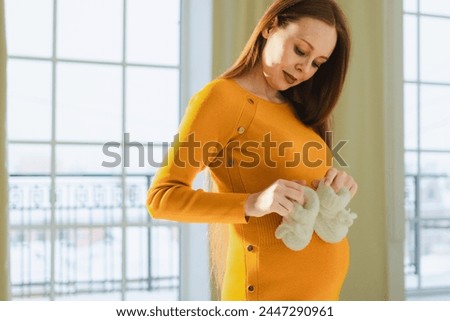 Pregnancy motherhood people expectation future. Pregnant woman with big belly holding newborn baby booties smiling at home. Young mom enjoying pregnancy. Maternity tenderness parenthood new life Royalty-Free Stock Photo #2447290961