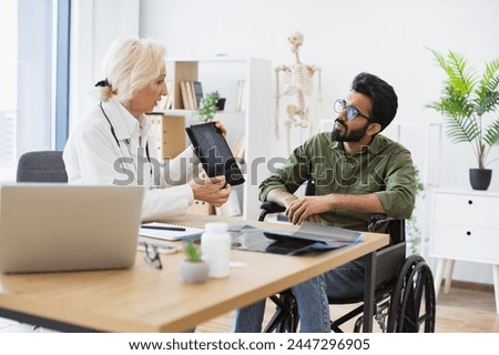 Efficient traumatologist reassuring patient about diagnosis in hospital interior. Senior lady in white coat demonstrating spine scans on tablet to mindful man using wheelchair.
