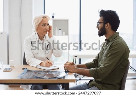 Mature elder physician giving recommendations and using notebook for writing prescription. Bearded gentleman describing symptoms of illness to senior female doctor and gesturing hands.
