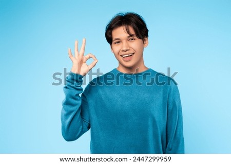 Cheerful young asian guy with a beaming smile showing an okay hand sign against a blue studio backdrop