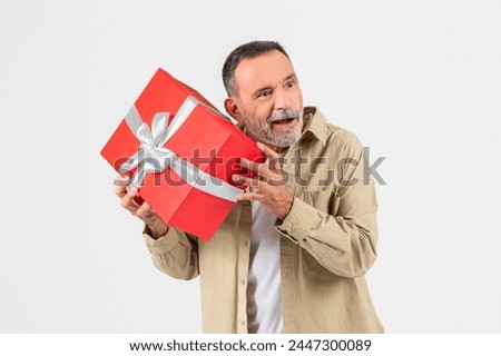 An older gentleman looks at a large red gift box curiously, suggesting anticipation or the act of giving a present