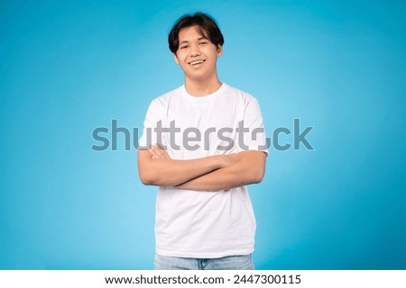 A smiling handsome young asian guy confidently poses with crossed arms against a lively blue backdrop Royalty-Free Stock Photo #2447300115