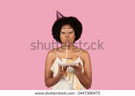 Surprised African American young lady blowing a candle on a piece of cake, wearing a birthday cap on pink