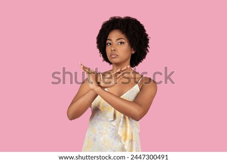 A young African American woman with curly hair in a floral dress stands against a pink background, crossing her hands in an X shape to signal stop or refusal Royalty-Free Stock Photo #2447300491