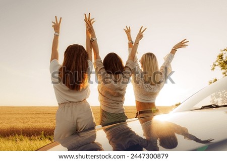Three joyful women raise their hands in celebration of freedom during a road trip at sunset, with a vast field in the background. Royalty-Free Stock Photo #2447300879