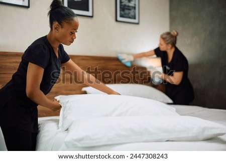Two maids cleaning guest room in a hotel. Focus is on African American maid.  Royalty-Free Stock Photo #2447308243