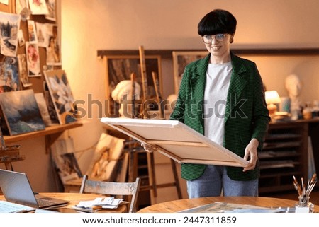 Waist up portrait of woman artist holding picture while standing in art studio or small business gallery, copy space