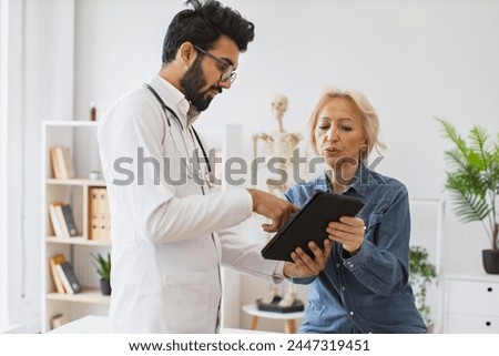 Young specialist writing instruction for taking medication correctly. Bearded male doctor and elderly female patient sitting at exam couch of modern medical center and diagnose disease.
