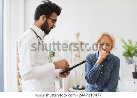 Respected man doctor and senior woman patient sitting at exam couch of new medical center and diagnose disease. Bearded specialist writing electronic instruction for taking medication correctly.