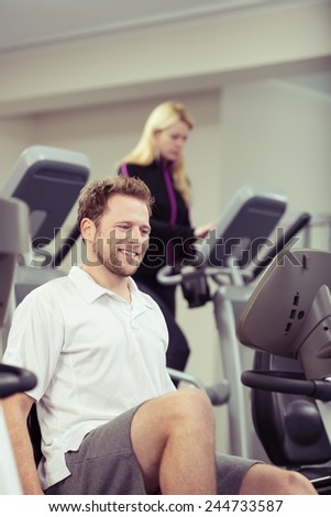 Smiling young man exercising on the equipment at the gym with a young woman working out behind in a healthy lifestyle concept