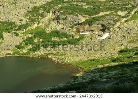 Top view of a fragment of a large lake surrounded by gentle slopes of high mountains with loose stones and sparse thickets of grass. Ergaki Natural Park, Krasnoyarsk Territory, Siberia, Russia. Royalty-Free Stock Photo #2447392013
