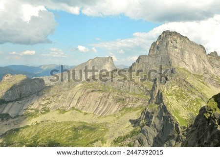 High and narrow ridges of jagged mountain peaks, partly covered with grass under a cloudy summer sky. Ergaki Natural Park, Krasnoyarsk Territory, Siberia, Russia. Royalty-Free Stock Photo #2447392015