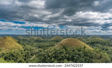Amazing karst rounded hills covered with brownish grass stretched to the horizon. Tropical vegetation all around. Clouds in the blue sky. Chocolate Hills Natural Monument. Philippines. Bohol. Royalty-Free Stock Photo #2447422537