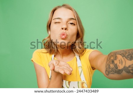 Close up young housewife housekeeper chef cook baker woman wears apron yellow t-shirt do selfie shot on mobile cell phone blow air kiss wink isolated on plain green background. Cooking food concept