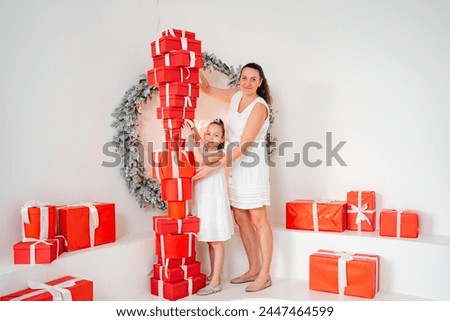 mom and little daughter in white dress on white wall background with red gift boxes and wreath. New Year's decor. happy childhood 